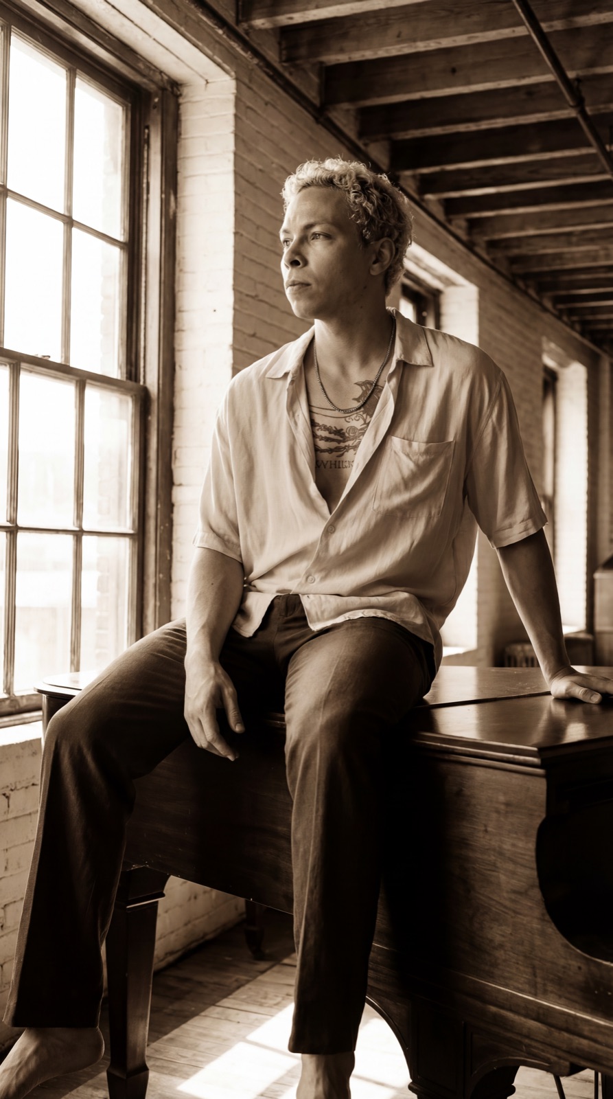 Kaelum seated on an old piano in warm light, looking towards the window